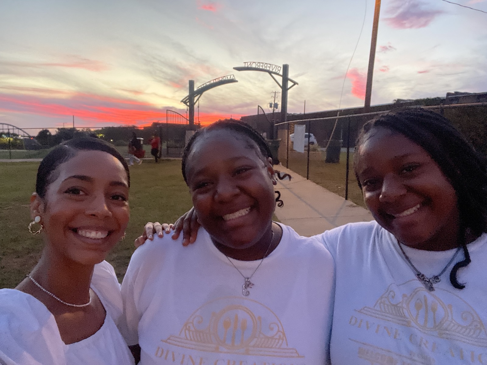 Ella and two young women smiling together at sunset on a riverside walkway in Selma